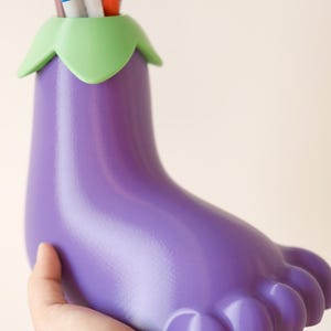 May include: Purple eggplant-shaped desk organizer with a green top, holding a variety of pens and markers.