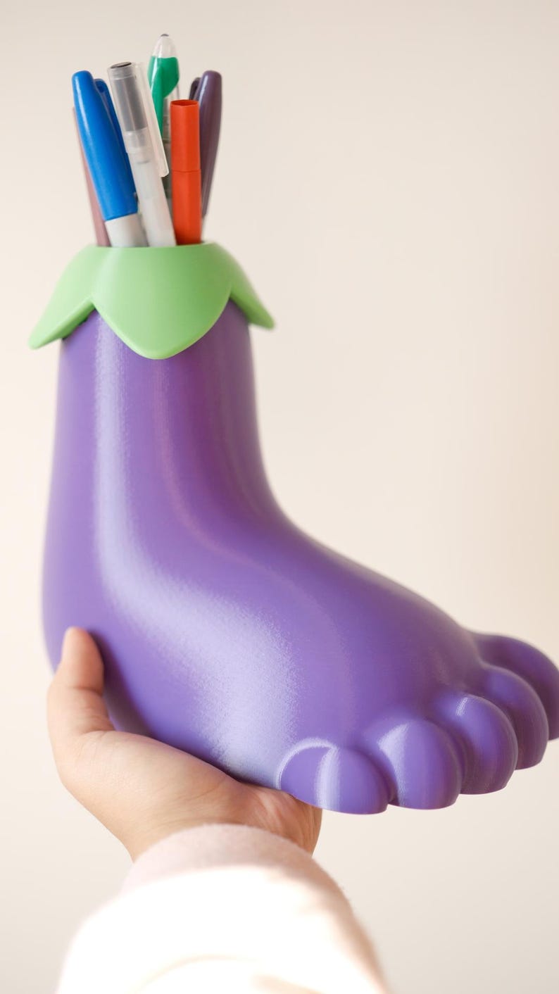 May include: Purple eggplant-shaped desk organizer with a green top, holding a variety of pens and markers.