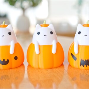 May include: Three Halloween-themed decorations featuring white cat figures emerging from orange pumpkin bases. The pumpkins have different carved faces, including a smiling face, a dot, and a jack-o'-lantern design. Each cat has black eyes and a small green stem.