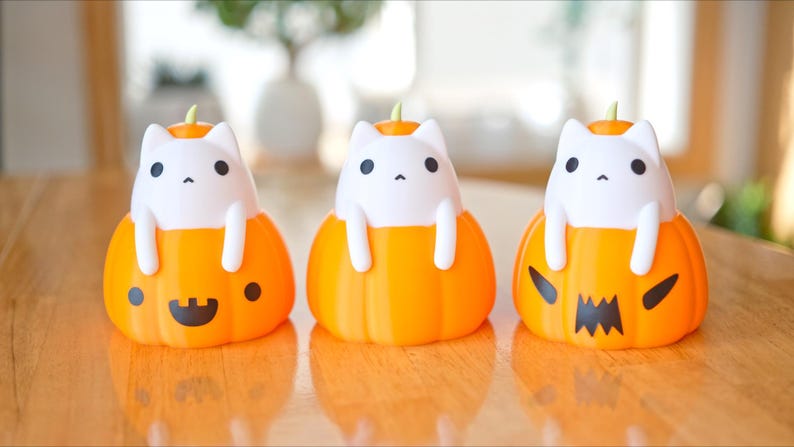 May include: Three Halloween-themed decorations featuring white cat figures emerging from orange pumpkin bases. The pumpkins have different carved faces, including a smiling face, a dot, and a jack-o'-lantern design. Each cat has black eyes and a small green stem.