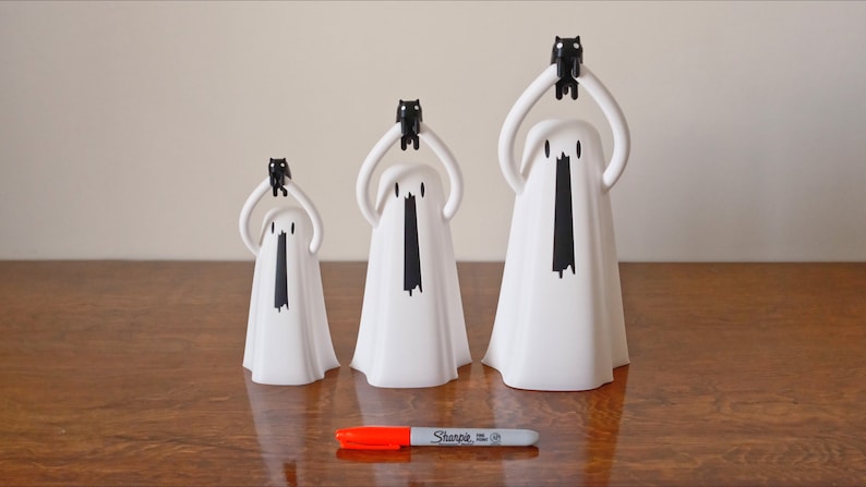 Peut inclure: Trois figurines de fant&ocirc;mes blanches avec des figurines de chats noirs sur la t&ecirc;te. Les fant&ocirc;mes sont de tailles diff&eacute;rentes et sont dispos&eacute;s par ordre de taille du plus petit au plus grand. Les fant&ocirc;mes ont des yeux et une bouche noirs. Les figurines sont sur une surface en bois.