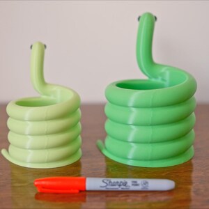 May include: Two coiled snake-shaped planters in light green and green, with small black eyes. A red and gray Sharpie marker is in front of the planters. The planters are made of a 3D-printed material.