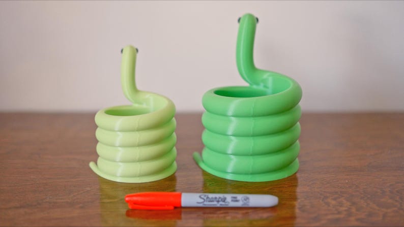 May include: Two coiled snake-shaped planters in light green and green, with small black eyes. A red and gray Sharpie marker is in front of the planters. The planters are made of a 3D-printed material.