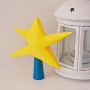 May include: A yellow star-shaped tree topper with a blue base. The star is made of paper and has a 3D effect. The base is a cylinder and is made of plastic.