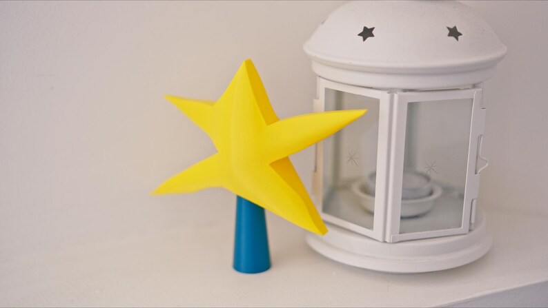 May include: A yellow star-shaped tree topper with a blue base. The star is made of paper and has a 3D effect. The base is a cylinder and is made of plastic.
