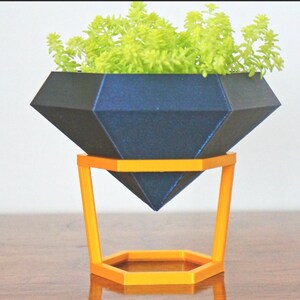 May include: A dark blue diamond-shaped planter with a green succulent plant inside, sitting on a gold metal stand with a hexagonal base.