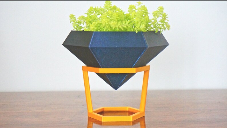 May include: A dark blue diamond-shaped planter with a green succulent plant inside, sitting on a gold metal stand with a hexagonal base.