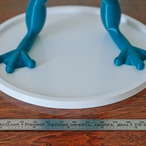 May include: A teal blue frog figurine with a white circular base. The frog has a textured surface and is standing on its hind legs. The base is approximately 4 inches in diameter.
