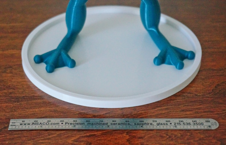 May include: A teal blue frog figurine with a white circular base. The frog has a textured surface and is standing on its hind legs. The base is approximately 4 inches in diameter.