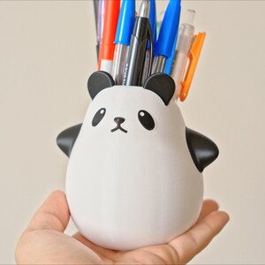 Op de afbeelding: Een witte, panda-vormige potloodhouder van keramiek met zwarte oren, ogen en neus. De houder is gevuld met een verscheidenheid aan pennen en potloden, waaronder blauwe, rode en zwarte.