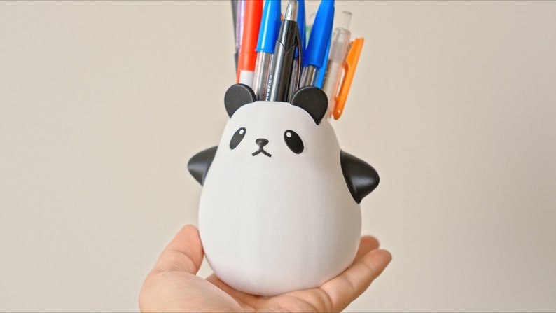 Op de afbeelding: Een witte, panda-vormige potloodhouder van keramiek met zwarte oren, ogen en neus. De houder is gevuld met een verscheidenheid aan pennen en potloden, waaronder blauwe, rode en zwarte.