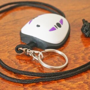 May include: A white and black key chain with a No-Face design, featuring purple accents. The key chain is attached to a black lanyard with a silver-colored key ring. The lanyard is knotted at the top.