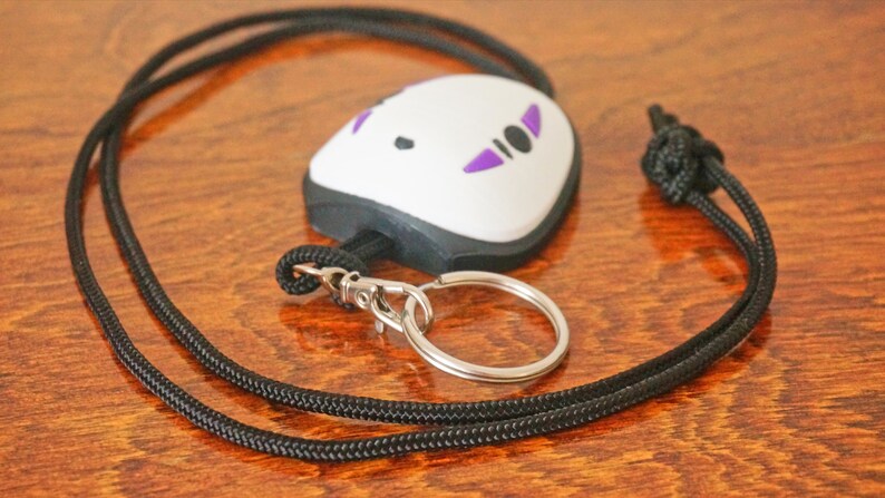 May include: A white and black key chain with a No-Face design, featuring purple accents. The key chain is attached to a black lanyard with a silver-colored key ring. The lanyard is knotted at the top.