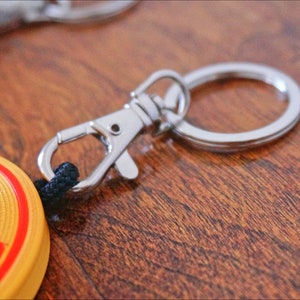 Puede incluir: Un llavero circular amarillo y rojo con un car&aacute;cter chino. El llavero tiene un anillo de metal plateado y un cierre de metal plateado.