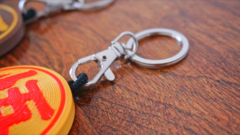 Puede incluir: Un llavero circular amarillo y rojo con un car&aacute;cter chino. El llavero tiene un anillo de metal plateado y un cierre de metal plateado.