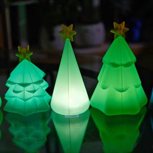 May include: Five illuminated Christmas tree decorations in varying shades of green and blue. Each tree has a yellow star on top. The trees are arranged in a row, reflecting on a dark surface.