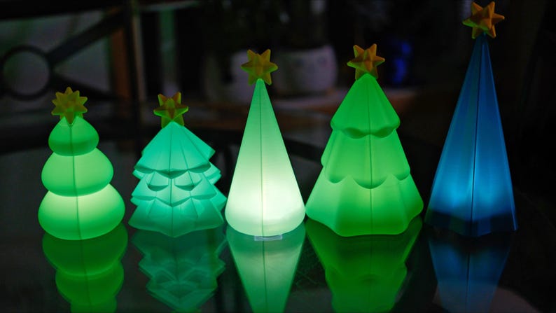 May include: Five illuminated Christmas tree decorations in varying shades of green and blue. Each tree has a yellow star on top. The trees are arranged in a row, reflecting on a dark surface.