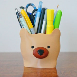 Puede incluir: Un porta l&aacute;pices con forma de oso marr&oacute;n con nariz y ojos negros. El porta l&aacute;pices est&aacute; lleno de varios bol&iacute;grafos, l&aacute;pices y marcadores, incluyendo amarillo, azul, verde y negro.