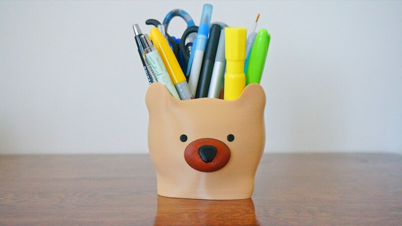 Puede incluir: Un porta l&aacute;pices con forma de oso marr&oacute;n con nariz y ojos negros. El porta l&aacute;pices est&aacute; lleno de varios bol&iacute;grafos, l&aacute;pices y marcadores, incluyendo amarillo, azul, verde y negro.