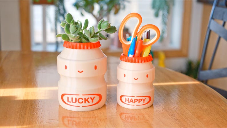 May include: Two cream-colored, smiling, bottle-shaped containers with orange lids. One holds a succulent plant, the other scissors and pens. The first reads "LUCKY" and the second "HAPPY" in orange. A cheerful desk organizer.