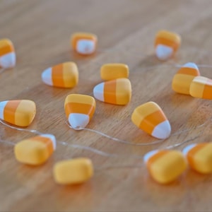 May include: A string of decorative candy corn lights featuring multiple small, triangular-shaped ornaments in yellow, orange, and white. The ornaments are connected by a thin, clear wire and include a small white battery pack at the end of the strand.