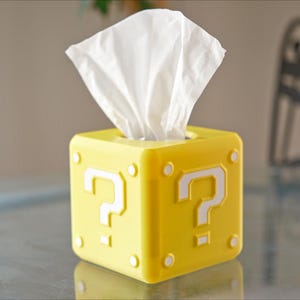 K&ouml;nnte beinhalten: Gelbe Tissuebox in Form eines Super Mario Bros. Frageblocks mit einem wei&szlig;en Fragezeichen auf jeder Seite. Die Box ist mit wei&szlig;en Taschent&uuml;chern gef&uuml;llt.