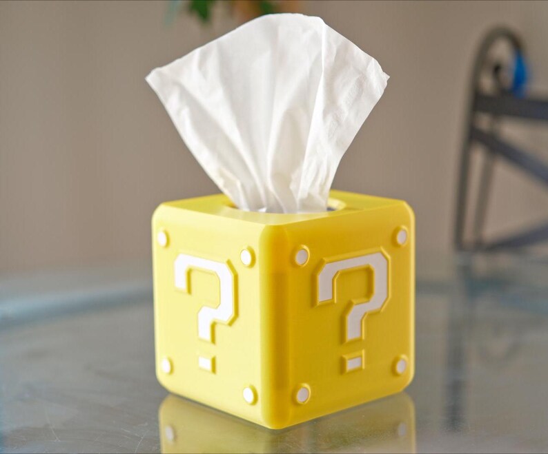 K&ouml;nnte beinhalten: Gelbe Tissuebox in Form eines Super Mario Bros. Frageblocks mit einem wei&szlig;en Fragezeichen auf jeder Seite. Die Box ist mit wei&szlig;en Taschent&uuml;chern gef&uuml;llt.