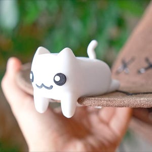 May include: A white cat ornament with black eyes and a smiling mouth sits on the brim of a brown hat. The hat features decorative stitching. The figurine is a charming, whimsical decoration.