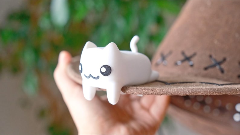 May include: A white cat ornament with black eyes and a smiling mouth sits on the brim of a brown hat. The hat features decorative stitching. The figurine is a charming, whimsical decoration.