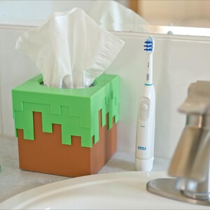 A cube tissue box cover inspired by the dirt grass block from the popular video game, Minecraft, is on a bathroom counter next to the sink and a bottle of soap. It has bright green pixelated grass at the top half and the bottom half is brown.