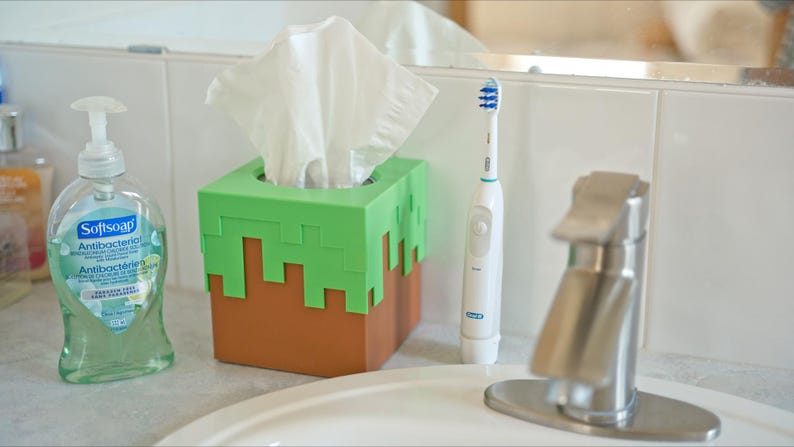 A cube tissue box cover inspired by the dirt grass block from the popular video game, Minecraft, is on a bathroom counter next to the sink and a bottle of soap. It has bright green pixelated grass at the top half and the bottom half is brown.