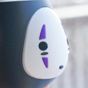 May include: A black and white ceramic cup featuring a stylized face with purple accents. The cup has a rounded shape and is held by a hand. The design is reminiscent of a character from an animated film.