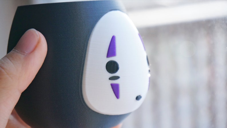May include: A black and white ceramic cup featuring a stylized face with purple accents. The cup has a rounded shape and is held by a hand. The design is reminiscent of a character from an animated film.