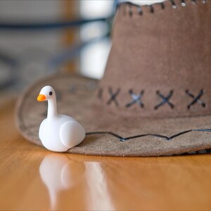 May include: A white figurine of a duck with an orange beak sits next to a brown cowboy hat. The hat has a stitched brim and decorative stitching. The items are on a wooden surface.