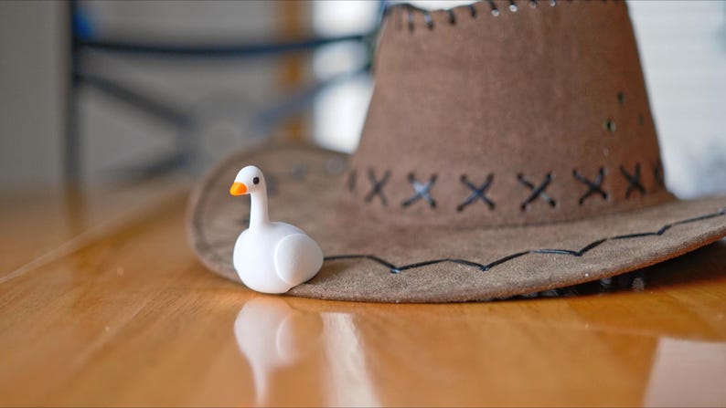 May include: A white figurine of a duck with an orange beak sits next to a brown cowboy hat. The hat has a stitched brim and decorative stitching. The items are on a wooden surface.