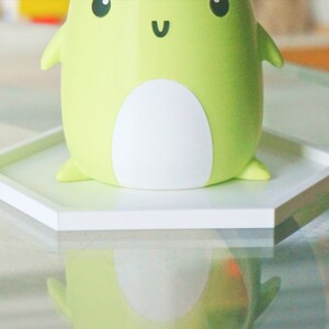 Puede incluir: Un posavasos hexagonal blanco con una figurita de rana verde en la parte superior. La rana tiene la barriga blanca y una cara feliz con ojos negros y una sonrisa.