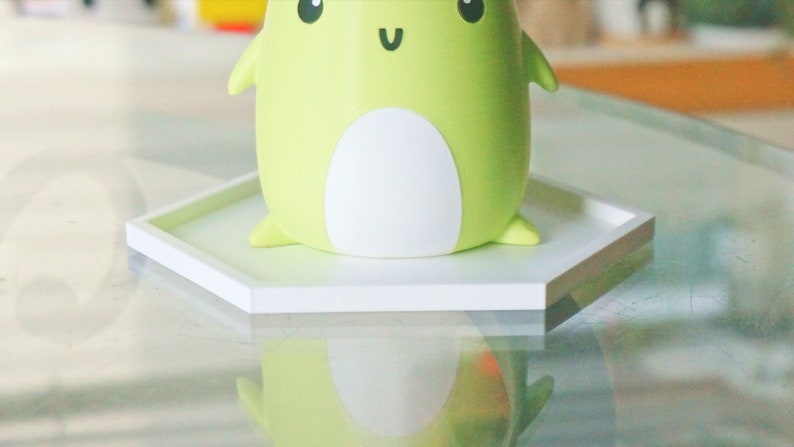 Puede incluir: Un posavasos hexagonal blanco con una figurita de rana verde en la parte superior. La rana tiene la barriga blanca y una cara feliz con ojos negros y una sonrisa.