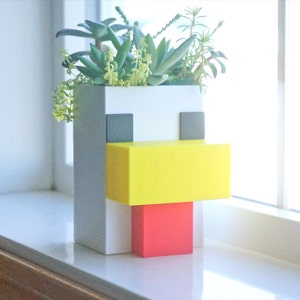 May include: A white, gray, yellow, and red planter shaped like a pixelated chicken with succulents growing inside.