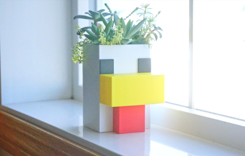 May include: A white, gray, yellow, and red planter shaped like a pixelated chicken with succulents growing inside.