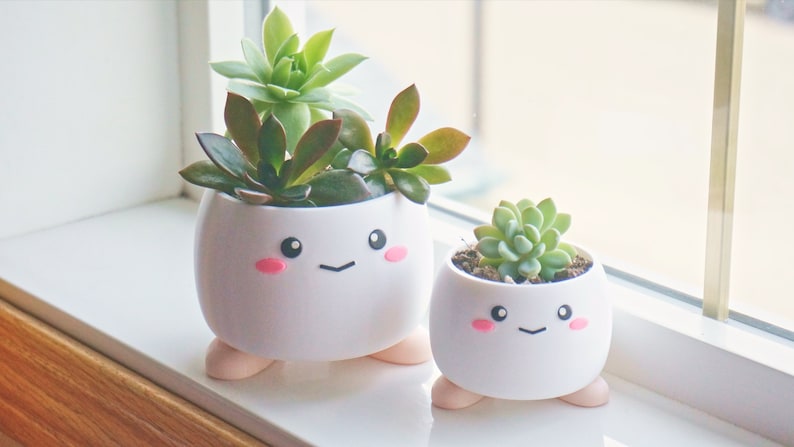May include: Two white ceramic planters with cute cartoon faces and pink cheeks. The planters have small legs and are filled with green succulents.