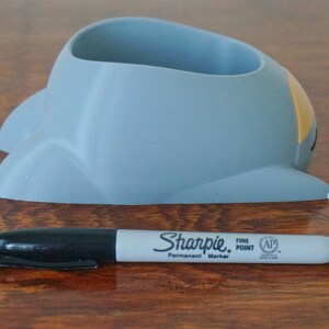 May include: A gray ceramic pencil holder shaped like a cartoon spaceship with a yellow accent on the side. The holder is sitting on a wooden surface.