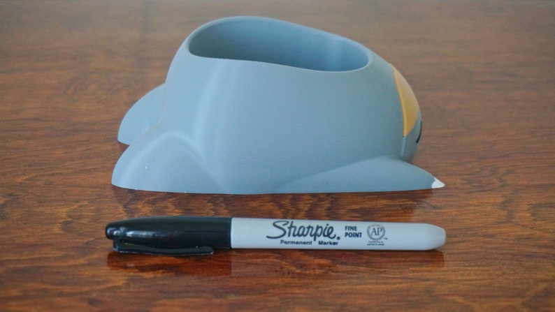 May include: A gray ceramic pencil holder shaped like a cartoon spaceship with a yellow accent on the side. The holder is sitting on a wooden surface.