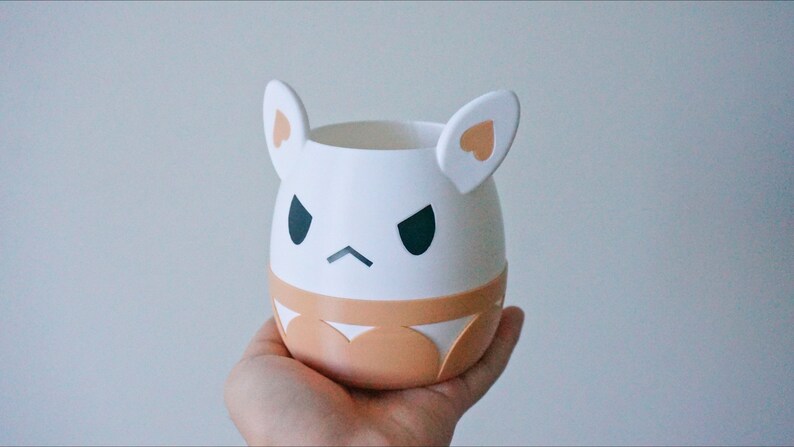 May include: White ceramic planter shaped like a cartoon animal with brown ears and a brown band around the middle. The animal has a grumpy expression with black eyes and a frown.