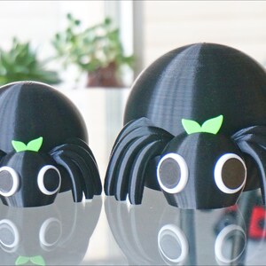 May include: Two black 3D printed spiders with googly eyes and green leaves on their heads. The larger spider is in the background and the smaller spider is in the foreground.