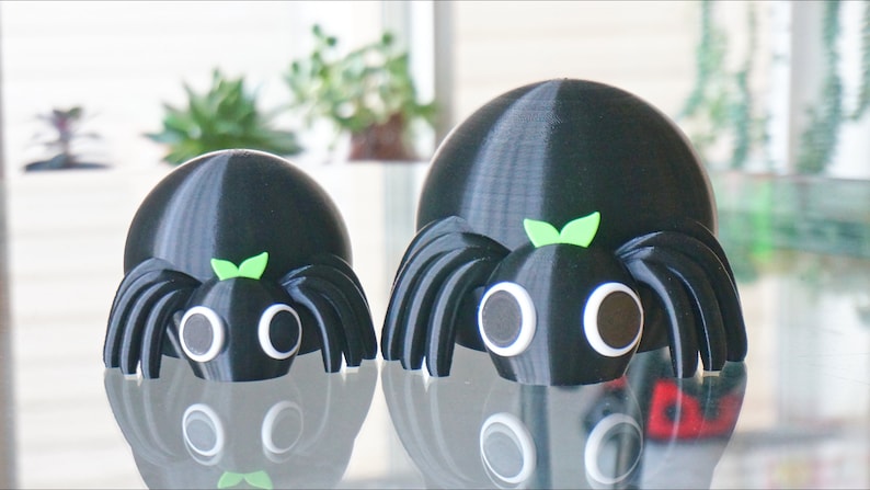 May include: Two black 3D printed spiders with googly eyes and green leaves on their heads. The larger spider is in the background and the smaller spider is in the foreground.