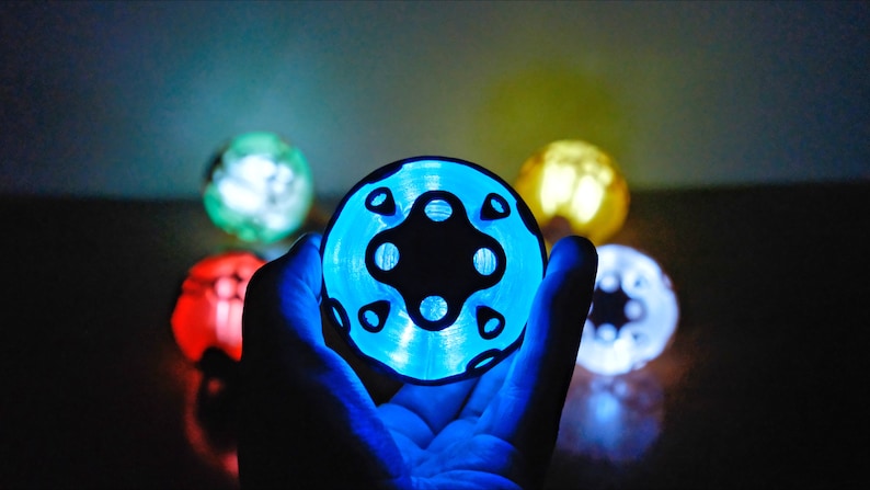 May include: A glowing, circular object with a black, symmetrical design is held in a hand. The object emits a bright blue light, with other glowing spheres in red, green, yellow, and white in the background. The overall effect is futuristic.