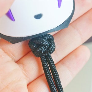 May include: A white and black charm with purple accents, featuring a character design. The charm is attached to a black knotted cord. The cord is held in a person's hand.