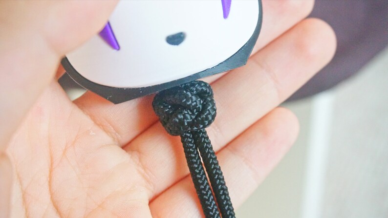 May include: A white and black charm with purple accents, featuring a character design. The charm is attached to a black knotted cord. The cord is held in a person's hand.