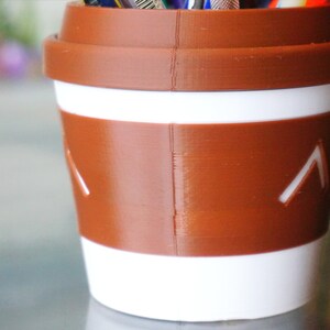 May include: A brown and white coffee cup shaped pencil holder with a white stripe and a brown band. The band has two white arrows pointing up.