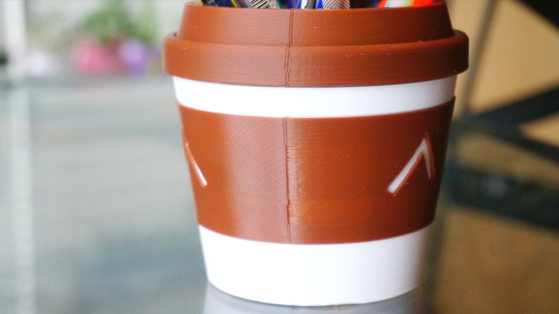 May include: A brown and white coffee cup shaped pencil holder with a white stripe and a brown band. The band has two white arrows pointing up.